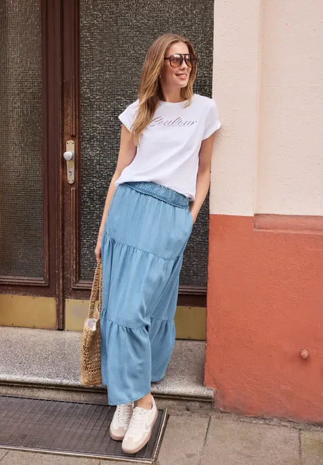 Junge Frau mit weißem T-Shirt, langem blauen Rock, Korbtasche und Sneakers lehnt an Hauswand.
